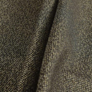 Puede incluir: Primer plano de una tela texturizada con un patr&oacute;n tejido. La tela tiene una mezcla de hilos gris oscuro, negros y beige, creando una apariencia moteada. El material est&aacute; doblado, mostrando profundidad y textura.