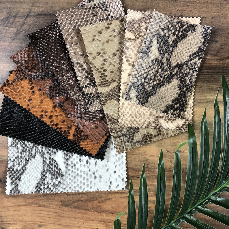 May include: Assortment of faux snakeskin fabric swatches in various colors and patterns, including brown, black, white, and beige. The swatches are arranged in a fan shape on a wooden surface.