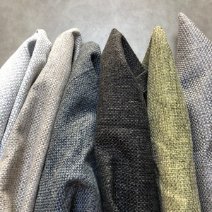 May include: Close-up of several rolls of textured fabric in various neutral colors. The fabrics include shades of gray, blue, and a dark charcoal, with a woven appearance. The folds and textures are visible, suggesting upholstery or drapery material.
