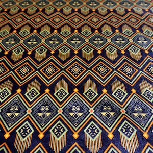 May include: Close-up of a fabric with a repeating geometric pattern. The design features diamond and rectangular shapes in shades of navy blue, orange, and light green. The pattern has a symmetrical, tribal-inspired aesthetic, creating a visually rich texture.