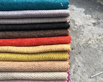 Pode incluir: Um conjunto de amostras de tecido em várias cores, incluindo turquesa, cinza, preto, marrom, laranja, amarelo, bege, branco, rosa e roxo. As amostras estão dobradas e empilhadas em uma superfície de concreto cinza.