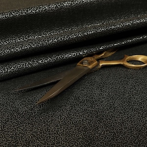 May include: A pair of gold-toned scissors with black handles, lying on a black fabric with a white swirl pattern.