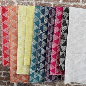 May include: A display of colorful fabric rolls, each showcasing a geometric triangle pattern with a speckled design. The fabrics vary in shades of red, orange, yellow, teal, navy, purple, pink, black, and gray, hanging against a brick wall.