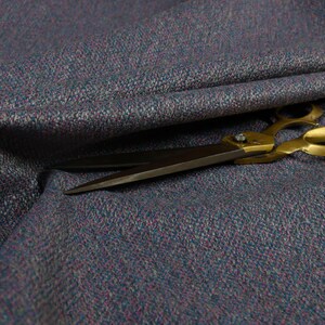 Puede incluir: Primer plano de una tela texturizada en tonos azules, morados y grises. Un par de tijeras con mango dorado descansa sobre la tela, con las hojas abiertas. La tela parece ser un material tejido, posiblemente lana o un textil similar.