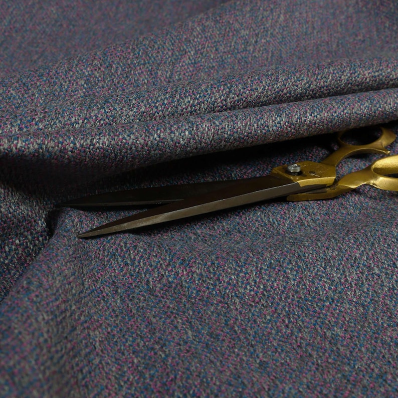 Puede incluir: Primer plano de una tela texturizada en tonos azules, morados y grises. Un par de tijeras con mango dorado descansa sobre la tela, con las hojas abiertas. La tela parece ser un material tejido, posiblemente lana o un textil similar.
