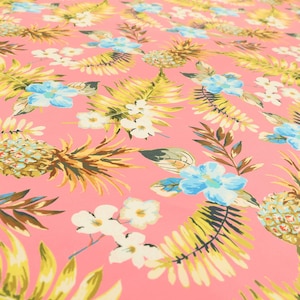 Op de afbeelding: Roze stof met een tropische print met ananas, bloemen en bladeren. De stof heeft een lichtroze achtergrond met gele, groene en blauwe accenten.