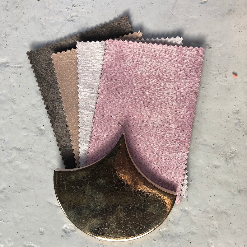 May include: A stack of fabric swatches in various shades of brown, gray, and pink. The swatches have a textured surface and are arranged in a fan shape. A gold-colored, crescent-shaped object is in the foreground.