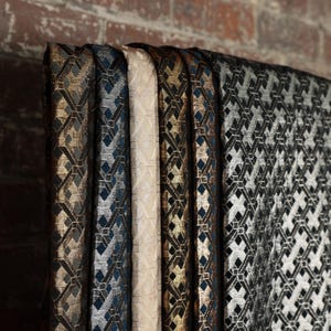 Pode incluir: Amostras de tecido estampado em várias cores, incluindo dourado, preto, prateado e creme. O tecido apresenta um design geométrico de diamante. As amostras estão penduradas contra uma parede de tijolos.