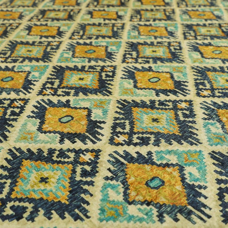 Op de afbeelding: Een beige kleed met een geometrisch patroon in tinten blauw, geel en oranje. Het patroon bestaat uit herhalende ruiten en vierkanten.