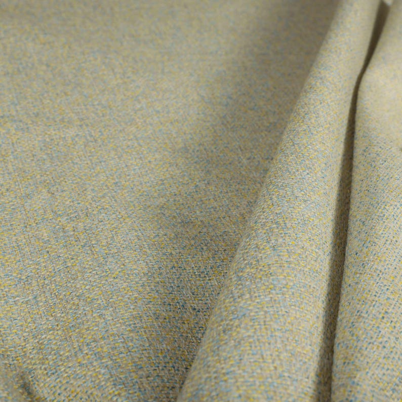 Puede incluir: Primer plano de una tela texturizada con un patr&oacute;n tejido. La tela es principalmente de color beige claro, con motas amarillas y azules. La tela est&aacute; doblada, mostrando la textura y la ca&iacute;da del material.