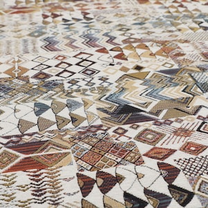 May include: A close-up of a fabric with a white background and a colourful geometric pattern. The pattern is made up of various shapes, including triangles, squares, and zigzags. The colours in the pattern include brown, red, blue, and gold.