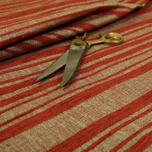 Op de afbeelding: Close-up van rood en beige gestreepte stof met een getextureerde weefsel. Een schaar met gouden handvatten ligt op de stof, de bladen open. De stof is opgevouwen en toont het patroon en de textuur.