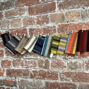 May include: Various fabric samples with striped patterns are pegged to a string against a red brick wall. The swatches showcase a range of colours, including black, grey, blue, green, orange, and burgundy, demonstrating diverse textures and designs for interior textiles.