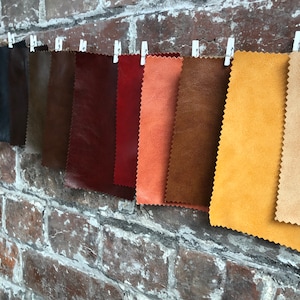 May include: A selection of leather fabric swatches in shades of black, brown, red, and tan, are hung against a brick wall. The samples are attached to a line with small white pegs, displaying the different textures and colours.