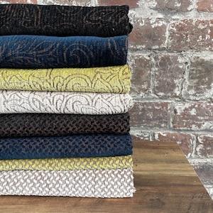 Könnte beinhalten: Ein Stapel von acht Stoffmustern in verschiedenen Farben und Mustern, darunter Schwarz, Marineblau, Braun, Grün und Weiß. Die Muster sind vertikal auf einer Holzoberfläche gestapelt.