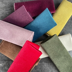 May include: A collection of fabric swatches in various colors, including red, teal, olive green, and yellow. The swatches are rectangular and have different textures, some with a velvet-like appearance and others with a woven texture. The edges are neatly finished.