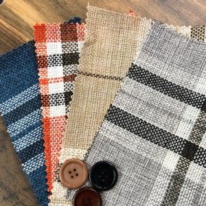 May include: Four fabric swatches in different patterns and colors. The swatches are arranged in a fan shape. The swatches are a light brown with a woven texture, a dark blue with a plaid pattern, a light gray with black stripes, and an orange and white plaid pattern. There are three buttons on the fabric swatches.