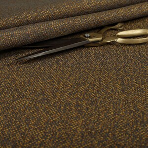 Puede incluir: Primer plano de tela de tweed marr&oacute;n y dorado con un tejido texturizado. Unas tijeras doradas descansan sobre la tela, con las hojas abiertas. La tela est&aacute; doblada en el fondo.