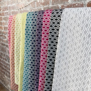May include: A selection of fabric rolls in a range of colours, including red, orange, yellow, teal, purple, magenta, black, and grey. Each roll features a white wavy pattern. The fabrics are displayed against a brick wall.