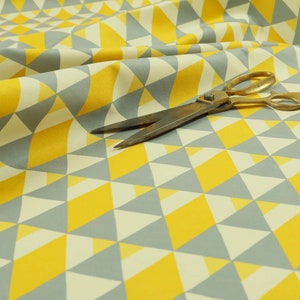 May include: A yellow and grey geometric fabric with a repeating pattern of triangles. A pair of scissors is lying on top of the fabric.
