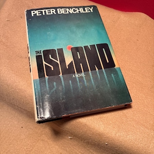 May include: A vintage paperback book titled "The Island" by Peter Benchley. The cover features a blue and orange gradient with the title in large black letters. The book is slightly worn, suggesting it is a used item.