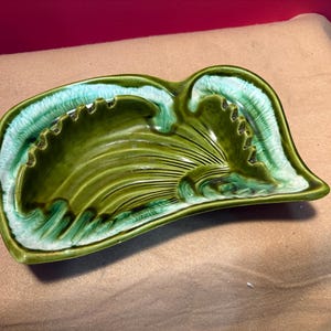 May include: A green ceramic ashtray with a leaf-like design. The ashtray has a wavy shape with a central depression and ridges. The edges are trimmed with a lighter green color. The ashtray is on a tan surface.
