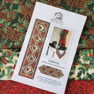 Puede incluir: Un patrón de papel blanco con un diseño de camino de mesa y cubrecama acolchado rojo y verde. El patrón se titula "Festivity" e incluye el texto "Quilting Time" y "Design by Vicki Fitratton".