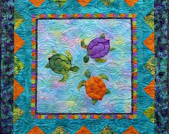 May include: A quilt with a blue background and a border of orange, green, and purple. Three colourful sea turtles are swimming in the centre of the quilt.