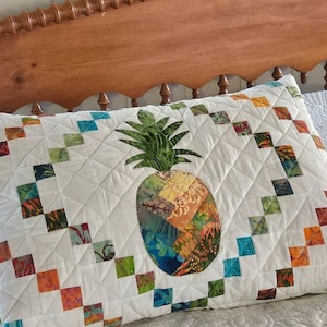 May include: A white pillow with a patchwork pineapple design in shades of green, blue, orange, and yellow. The pillow is on a white bed with a wooden headboard.