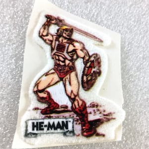 May include: A vintage sticker featuring He-Man, a muscular character from the Masters of the Universe franchise. He-Man is depicted in a dynamic pose, holding a sword and shield. The sticker has a white border and the text "HE-MAN".