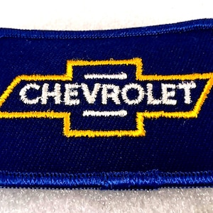 May include: A rectangular blue patch with a yellow and white Chevrolet logo. The logo features a stylized bow tie design with the word "CHEVROLET" in white. The patch has a blue stitched border.