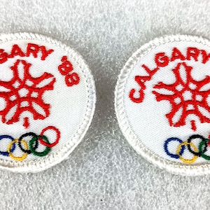 May include: Two circular white embroidered patches featuring the red text CALGARY '88 arched over a red stylized snowflake emblem. Below the snowflake, the five interlocking Olympic rings are embroidered in blue, yellow, black, green, and red.