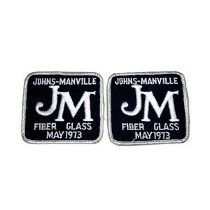 May include: Two square embroidered patches with black backgrounds and white borders. Each patch features the white text JOHNS-MANVILLE at the top, a large white JM monogram in the center, and the white text FIBER GLASS MAY 1973 at the bottom.