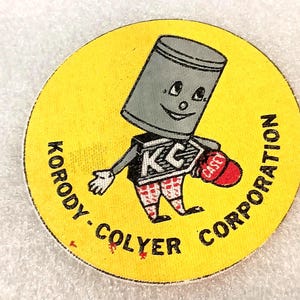 May include: A vintage yellow Korody-Colyer Corporation sticker. The sticker features a cartoon character with a gray cylinder head, a smiling face, and red and white plaid shorts. The character is holding a red baseball cap.