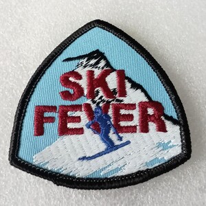May include: A triangular embroidered patch with a black border, featuring a light blue sky and a white snow-covered mountain. The text "SKI FEVER" is stitched in bold red letters, with a small blue silhouette of a person skiing down the slope.