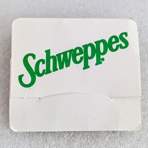 May include: A white, rectangular matchbook with the word "Schweppes" printed in green, stylized lettering. The matchbook has a perforated edge for easy tearing.
