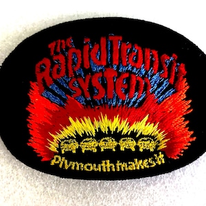 May include: An oval-shaped black embroidered patch featuring red and blue text that reads The Rapid Transit System. Below the text is a vibrant red and yellow flame design with five small yellow car icons and the text Plymouth makes it.