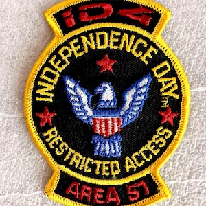 May include: Embroidered patch with a black background and yellow border. The patch features an eagle with blue and white wings and red and white stripes. Text includes "INDEPENDENCE DAY", "RESTRICTED ACCESS", and "AREA 51".