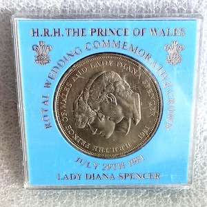 May include: A commemorative coin in a blue case. The coin features profiles of Prince Charles and Lady Diana Spencer, with text that reads "Royal Wedding Commemorative Crown" and "July 29th 1981".