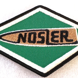May include: Embroidered patch with a green diamond shape, featuring the word "NOSLER" in black letters over a brown oval. The patch has a white border and a black outline.