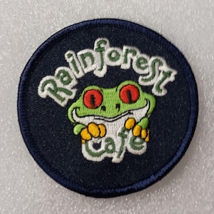 May include: A round, navy blue patch featuring a green frog with red eyes and yellow hands. The words "Rainforest Cafe" are embroidered in white and green, creating a whimsical design.