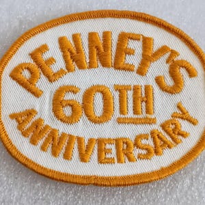 May include: An embroidered patch with an oval shape, featuring the text "PENNEY'S 60TH ANNIVERSARY" in orange thread on a white background. The patch has an orange border.