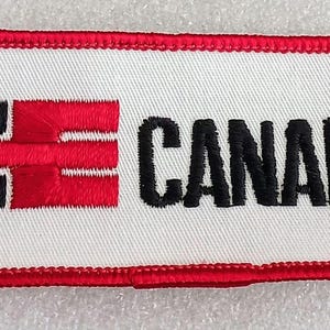 May include: Embroidered patch with a white background, red border, and the word "CANADA" in black. The patch features a stylized logo in black and red.