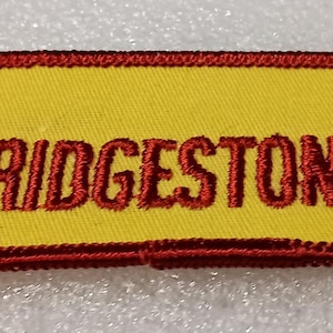 May include: Yellow rectangular patch with red embroidered text and border. The text reads "BRIDGESTONE TIRE" with a red logo of "BS" inside a red outline. The patch is likely for a uniform or apparel.