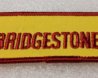 May include: Yellow rectangular patch with red embroidered text and border. The text reads "BRIDGESTONE TIRE" with a red logo of "BS" inside a red outline. The patch is likely for a uniform or apparel.