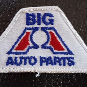 May include: White triangular patch with the words "BIG AUTO PARTS" in blue lettering. The design includes a stylized "A" in red and blue. The patch has a white border and is likely an emblem.