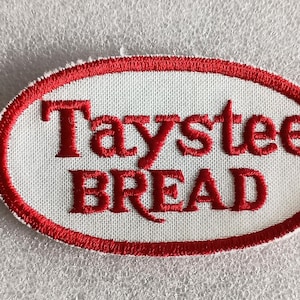 May include: Oval-shaped patch with the words "Taystee BREAD" embroidered in red on a white background. The patch has a red border and is likely for clothing or accessories.
