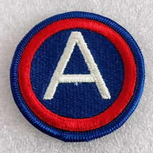 May include: A circular patch with a blue background, a red ring, and a white letter "A" in the center. The patch has a stitched border and is likely an emblem or insignia.
