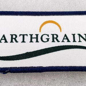 May include: A rectangular patch with a white background and a navy blue border. The text "EARTHGRAINS" is in dark green, with a gold semi-circle above it and a green curved line below. The patch is likely for apparel.