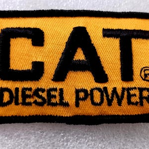 May include: Embroidered patch with a yellow background and black border. The patch features the word "CAT" in bold black letters above the words "DIESEL POWER". A registered trademark symbol is visible.